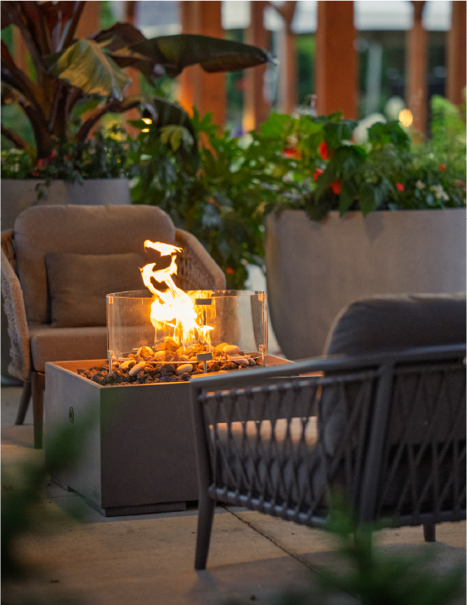 Outdoor patio with cushioned chairs around a glass-enclosed fire pit, surrounded by large potted plants and wooden beams. Outdoor patio with cushioned chairs around a glass-enclosed fire pit, surrounded by large potted plants and wooden beams.